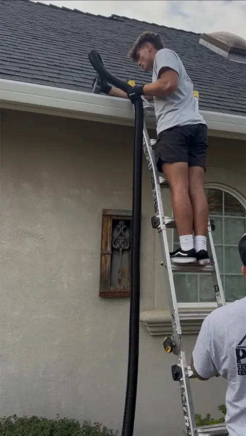 A crew member on a ladder feeding a large vacuum hose into a roof gutter while a second crew member steadies the ladder