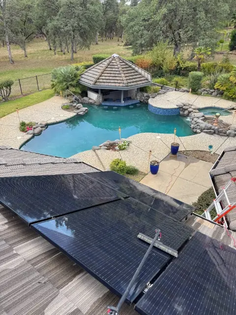 A brush on a long pole cleaning wet rooftop solar panels above a backyard pool