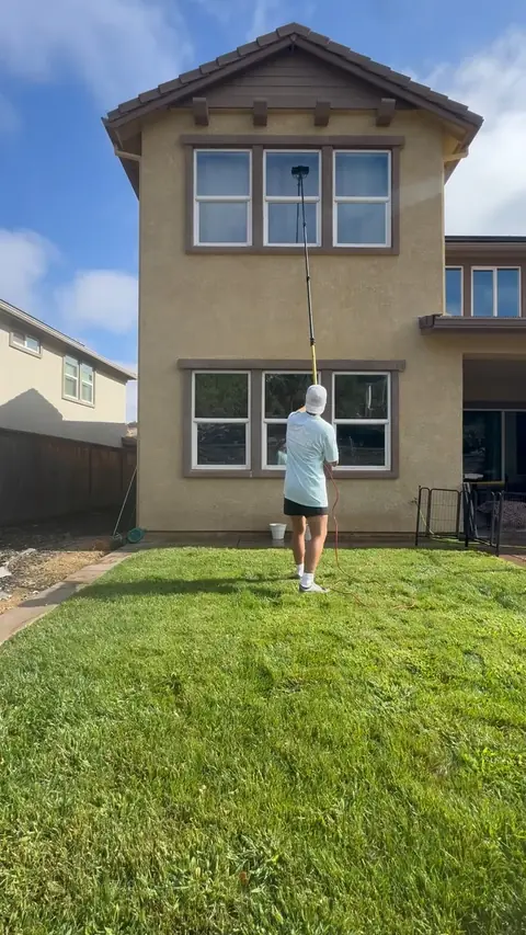 A crew member on a backyard lawn washing the second-story windows of a two-story house with a long pole