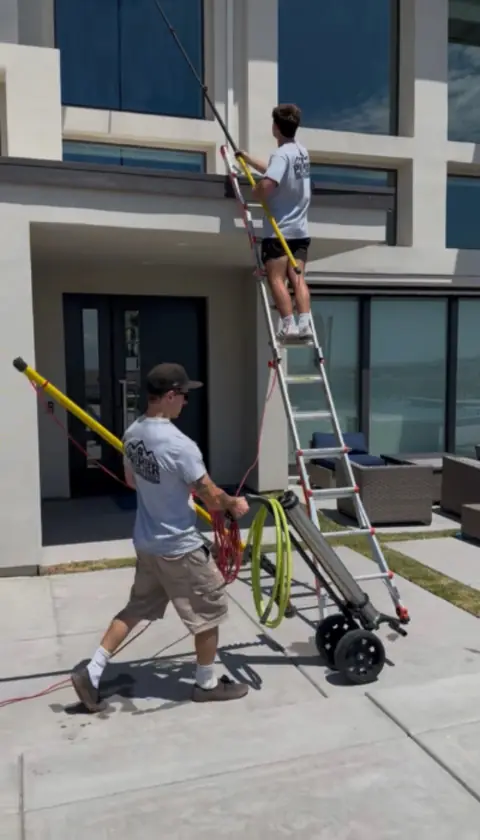Two Premier Exterior crew members at a modern two-story home, one on a ladder washing upper windows with a long pole, the other with a cart carrying a tank and hose reel