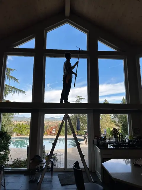 A crew member on a high interior ledge running a squeegee on a pole across a tall wall of windows