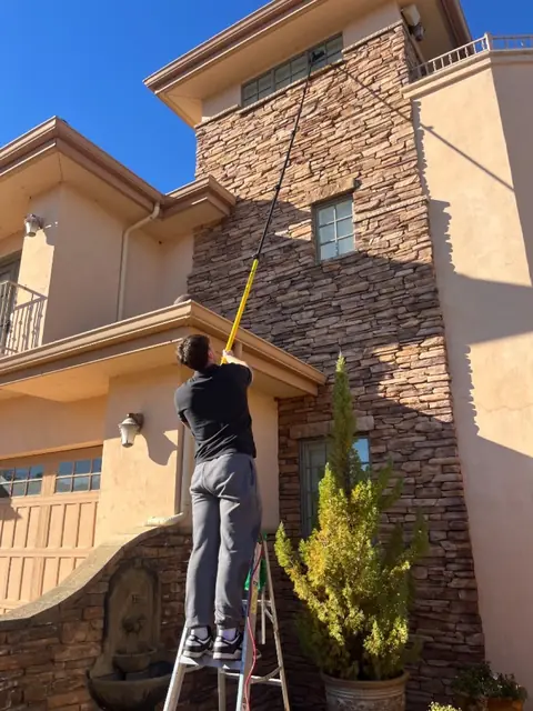 A crew member on a stepladder reaching a long pole up to a high window on a stacked-stone tower