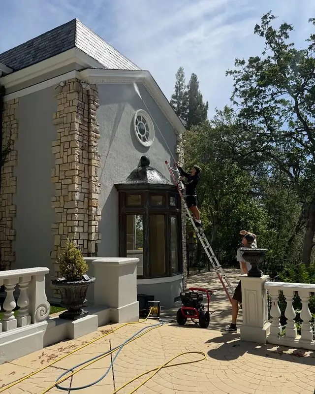 Two crew members washing a painted house wall, one spraying from a ladder and one on the patio below
