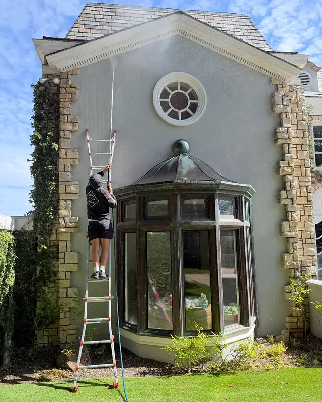 A crew member on a ladder spraying a tall painted gable above a bay window