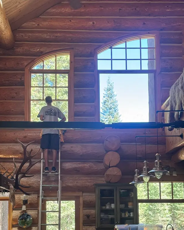 A crew member on a tall ladder cleaning the upper arched windows inside a log home
